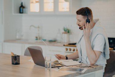 Modern mutfaktaki dizüstü bilgisayarla masada oturan ve müşteriyle online görüşme yaparken ekrana bakan odaklanmış sakallı çağrı yöneticisinin ya da serbest çalışanın yan görüntüsü. Serbest konsept