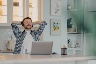 Happy young male worker resting after video call with colleagues and feeling relaxed after hard working day at home, using laptop and headset. Remote work and freelance concept