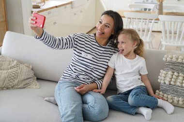 Funny little daughter and young mother showing tongue taking pictures of family, looking at mobile phone camera, happy mom having fun with kid girl, taking selfie together, sitting on couch at home.