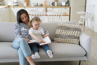 Anaokulu kızıyla birlikte kitap okuyor, öğreniyor, aile hobisinden zevk alıyor. Anne kucaklıyor, kızı eğitiyor, kanepede oturuyor, evde masal okuyor. Ebeveynlik, annelik..