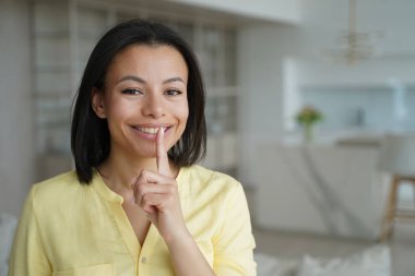 Woman gestures hush and smiling. Conceptual portrait of young positive emotional woman. Advertising banner mockup. Handsome european girl in living room at home. Concept of secret information.