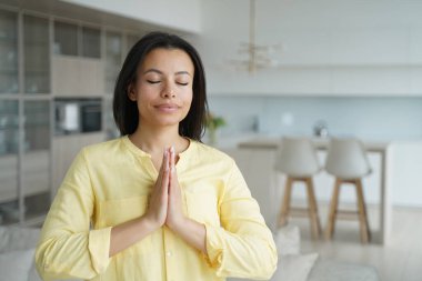 Calm young woman folded hands in namaste gesture, praying with closed eyes at home. Female practice yoga, meditating, breathing, relax her mind. Healthy lifestyle, anti-stress meditation.