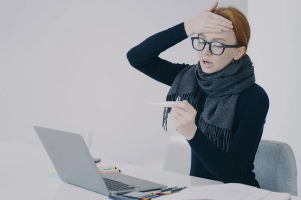 Sick manager at online conference. Young white woman got disease in office. Unwell girl is working in scarf holding thermometer and shocked with her temperature. Worker feels ill and has fever.