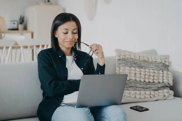 Confident sophisticated arab woman at work, career concept. Businesswoman is working from home. Lady is sitting in comfortable couch with laptop and smiling, looking at the screen.