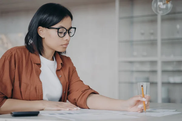 Serious woman in glasses calculates monthly expenses, taxes sitting at workplace desk in office. Focused businesswoman freelancer calculating finance, money, working on business project.