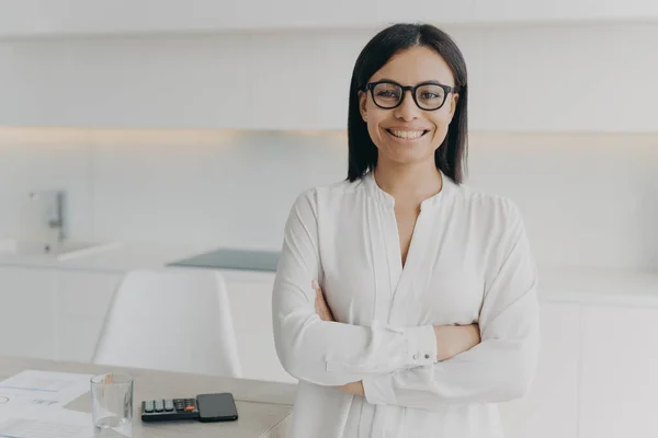 Young european executive or ceo in glasses and white blouse is working remote from home. Confident girl, entrepreneur is standing with her arms crossed and smiling. Career and leadership concept.