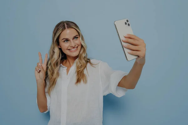 stock image Young happy woman in casual clothes taking selfie while showing peace gesture with her hand, smiling with toothy smile for followers and expressing herself, isolated over blue wall