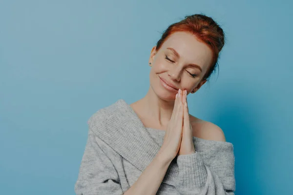 Happy calm redhead grateful woman holding hands in prayer gesture with closed eyes and tilted head, waiting for miracle with smile on face isolated on white blue studio background. Faith and hope