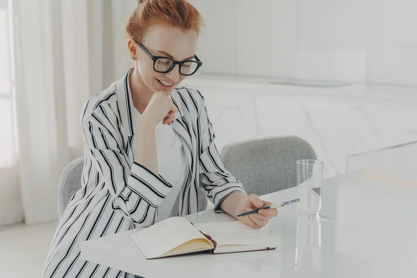 Indoor image of happy ginger business woman dressed casually sitting at table in kitchen, making notes and drinking pure water from glass in morning while planning her day, writing down thoughts ideas