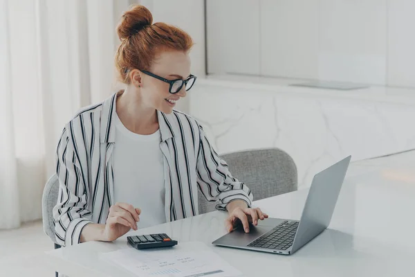 Young positive red-haired woman accounter or financier working remotely on laptop computer and using calculator while sitting at her home office, smiling while looking at pc screen, dressed casually