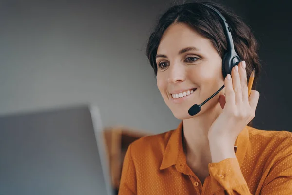 Close up of pleasant woman Spanish speaking call center employee in wireless headset with mic, smiling operator talking with customer online, looking at laptop screen while working remotely from home
