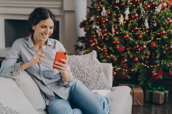 ワイヤレスイヤホンの幸せな女性は 明るい色のクリスマスツリーと自宅のリビングルームで居心地の良いソファに座って クリスマス時にオンラインで家族や友人と話して 手にスマートフォンを保持しています — ストック写真