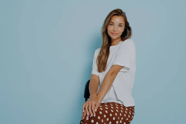 Beautiful caucasian model female sitting on chair with her hands folded on legs with positive face expression, wears casual white tshirt and brown polka dot skirt isolated on blue background