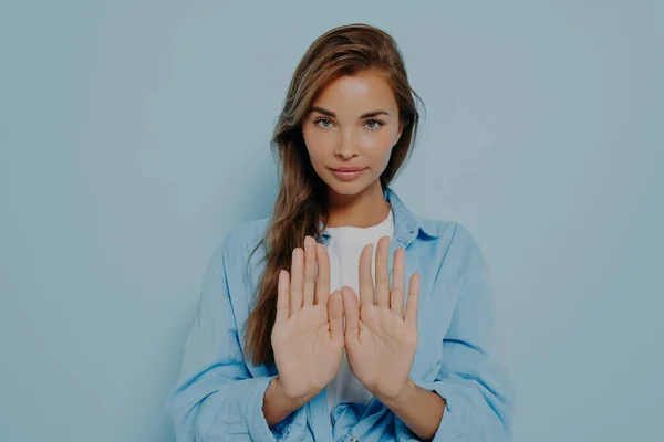 Long haired serious young woman showing stop gesture, asking to step back, protecting herself from unwanted attention or refusing to do something on light blue background