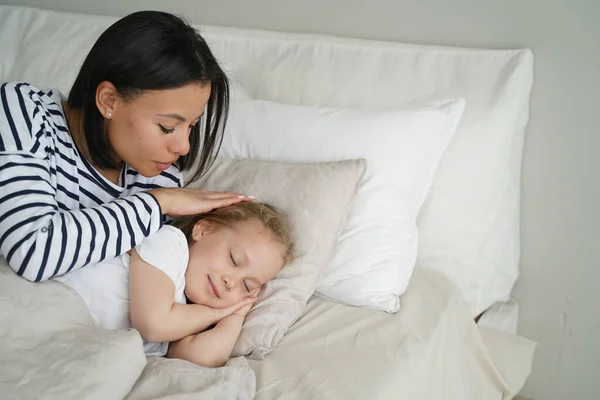 stock image Mom puts daughter to bed, stroke head of small child during daytime nap. Calm kid girl rest sleeping lying on soft pillow under fresh duvet. Healthy sleep in children, childcare.