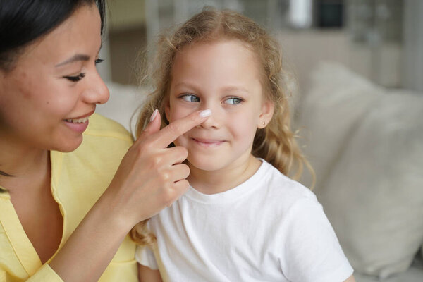 Любящая мать трогает нос маленькой приемной дочери. Улыбающаяся женщина чувствует любовь к маленькой девочке, наслаждается нежным моментом вместе дома. Счастливое материнство, уход за детьми, усыновление детей.