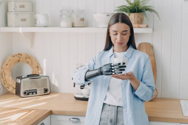 Pretty disabled girl is holding cup of tea with cyber hand carefully. Concept of grasp sensors in modern bionic prosthesis. Routine of handicapped person and life quality. Woman at domestic kitchen.