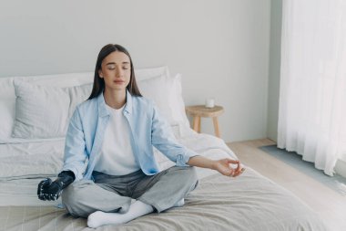 Meditation and concentration. Morning routine of disabled woman. Girl is sitting in lotus pose in her bedroom. Attractive european woman has artificial cyber limb. Stress relief at home.