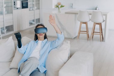 Caucasian handicapped girl in vr glasses. Disabled person gets rehabilitation getting used to sensor cyber arm. Young woman sitting in living room on couch. Amputee with myoelectric robotic limb.