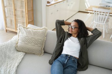 Calm african american young woman sitting relaxing on cozy sofa at home, holding hands behind head. Relaxed biracial girl dreaming, enjoying new house, resting on comfortable sofa in living room.