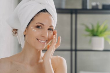 Face of beautiful european lady which applies cream. Attractive feminine caucasian girl wrapped in towel after bathing and hair washing. Happy young woman takes shower at home. Beauty routine.