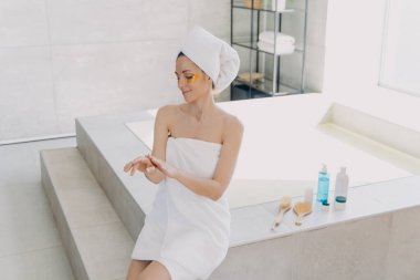 Girl applies eye patches and hand cream. Attractive caucasian woman wrapped in towel after bathing. Young hispanic lady takes shower at home. Relaxation at spa resort. Modern interior of bathroom.