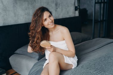 Gorgeous european woman combs her soft curly hair with hairbrush. Girl wrapped in towel doing hair treatment after bathing. Young woman takes shower at home and doing hair. Weekend morning at home.