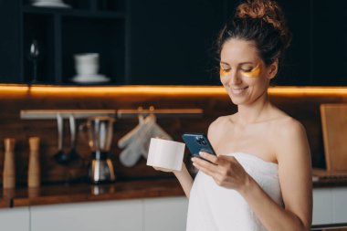 Lady relaxing at home, chatting on phone and having tea. Happy hispanic girl wrapped in towel after spa procedures. Young woman applies anti wrinkle eye patches. Body care, beauty and wellness.
