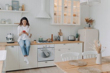 Mutlu genç bayanın telefonda mesajı var. Avrupalı ev hanımlarının boş vakitleri var ve evinde mutfaktaki atölyesinde oturuyorlar. Evde günlük kıyafetli bir kız. Modern lüks İskandinav iç mimarisi.