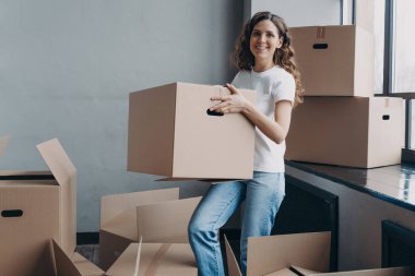 Girl in jeans and white t-shirt is carrying cardboard box. Happy attractive european woman packing things to move. Packed boxes on the floor. Relocation and moving to new apartment concept.