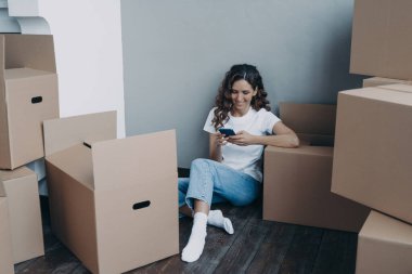 Kız cep telefonunda mesajlaşıyor ve yerde oturuyor. Karton paketler ve kutularla çevrili mutlu, çekici İspanyol kadın. Yeni daireye taşınmak kolay. Nakliye hizmeti konsepti.