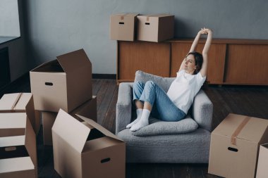 Lady is relaxing among boxes in living room of new home. Happy woman is an apartment buyer. Satisfied european girl is sitting in armchair. Dream house purchase and mortgage concept.