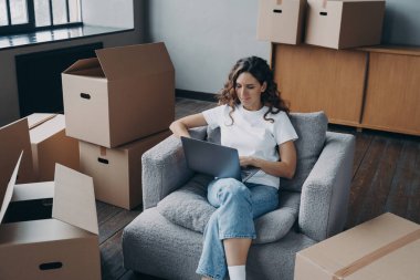 Happy young hispanic woman renting apartment online. Girl among cardboard boxes is using laptop and smiling. Happy homeowner is sitting in armchair with pc. Woman about to move.