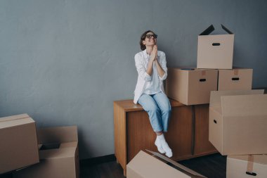 Happy young european woman purchases real estate and thanks god. Girl is sitting on boxes in new apartment and dreaming. Mover is excited. Concept of success and opportunities for women.