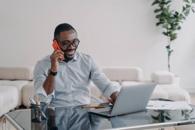 Freelancer is working online and having conversation with client on phone. Happy afro businessman has online meeting. Workplace at home office. Remote african american worker.
