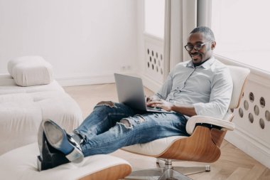 Smiling african american man uses laptop sitting in comfortable chair, black businessman looking at computer screen, communicating on social network, shopping online, working remotely at home.