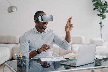 VR gözlüklü heyecanlı afro adam iş yerinde. İş adamının sanal toplantısı var. Freelancer tasarım projesi üzerinde çalışıyor. Uzak çalışma ve eğlence için modern kablosuz teknoloji.