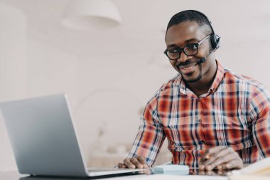 Smiling african american employee wearing headset working at laptop, looking at screen, answering a video call, communicates with customer. Black guy call center operator consultanting client.