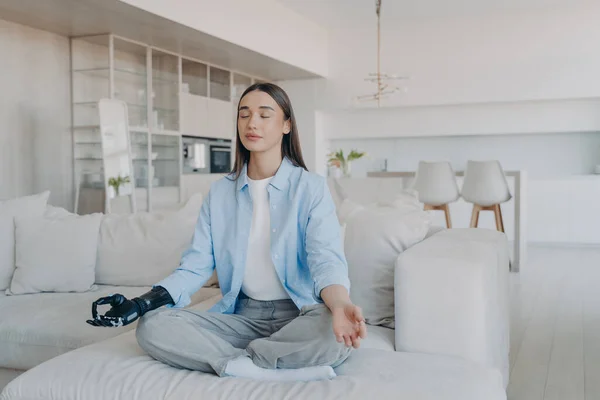 Morning yoga of disabled woman. Lady sitting in lotus pose on couch with her eyes closed. Attractive european girl has myoelectric cyber prosthesis. Modern artificial limb with nervous sensor control.