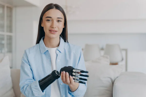 Happy disabled young woman is assembling bionic limb prosthesis. Attractive caucasian girl has cyber sensor hand. Female amputee setting her myoelectric arm at home. Innovations in medicine.