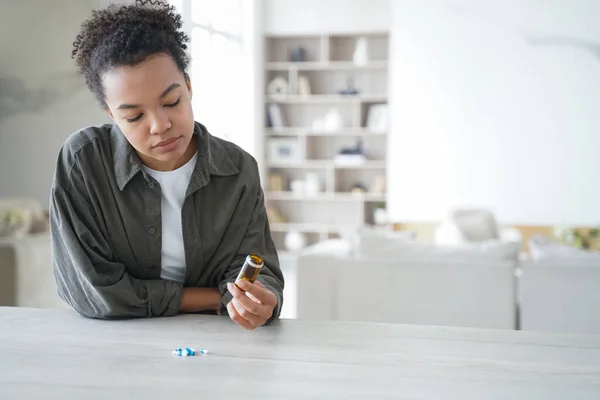 Unwell african american girl is holding pills flacon. Sick teenage girl is taking vitamins or painkillers. Young woman is upset and worried. Stressed patient. Pharmaceutical treatment concept.