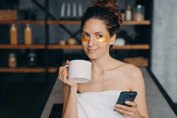 European girl drinks coffee and texting on smartphone. Young woman applies anti wrinkle eye patches and relaxing. Happy hispanic girl wrapped in towel after bathing. Evening body care routine.