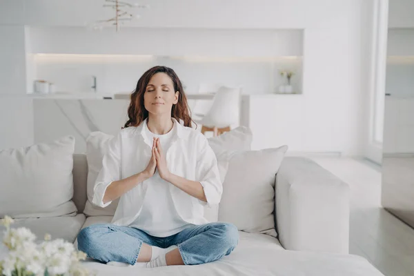 Serene latin woman at home. Posture exercise, lotus asana. Young european woman is practicing yoga and meditation on couch with her legs crossed. Minimal white modern scandinavian interior.