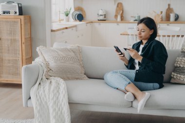 Kadın cep telefonu uygulaması ile banka hesabını kontrol ediyor, evdeki kanepede oturuyor. Elinde bankacılık kartı olan odaklanmış genç bir kadın. Akıllı telefon. Nakitsiz ödeme yapıyor. E-ticaret, çevrimiçi bankacılık kavramı.