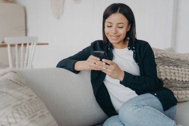 Akıllı telefonuyla koltukta oturan gülümseyen kadın. Elinde telefon tutan genç bir kadın, sosyal ağlardan arkadaşlarıyla haber mesajları paylaşıyor, internet hizmetlerinden hoşlanıyor, evde online alışveriş yapıyor..