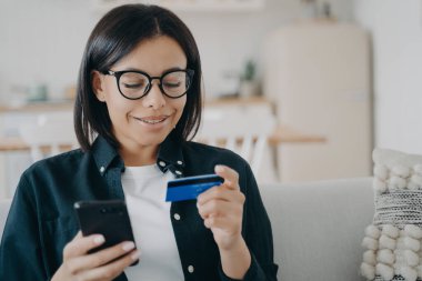 Smiling young woman in glasses holding bank credit card, smartphone in hands, making payment in shopping application, purchasing in online store at home. E-commerce, e-bank, online banking.