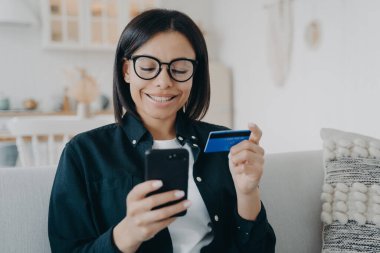 Smiling female holding bank credit card and smartphone. Young woman pays for purchases in online store using banking service, shopping, making secure payment, sitting on sofa at home. E-commerce.