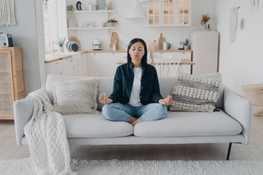 Huzurlu kadın yoga yapıyor, Mudra jestleriyle, oturma odasında kanepede oturarak nefes alıyor. Genç bayan meditasyon yapıyor, stresten kurtuluyor, evde dinleniyor. Sağlıklı yaşam tarzı, duygu yönetimi.