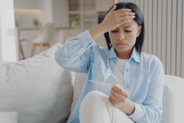 Uykusuz kadın ateşi ölçüyor ve kafaya dokunuyor. Avrupalı kız evdeki kanepede oturuyor ve elinde termometre var. Virüs enfeksiyonu ve gripten dolayı yorgunluk. Hastalık izni kavramı.