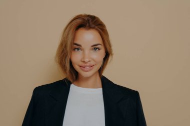 Beyaz bluz üzerine giyilen resmi siyah ceketli, kendine güvenen bir iş kadını portresi. Stüdyoda, boş bir alanda bej arka planda yalnız poz veriyor. Genç kadın model.